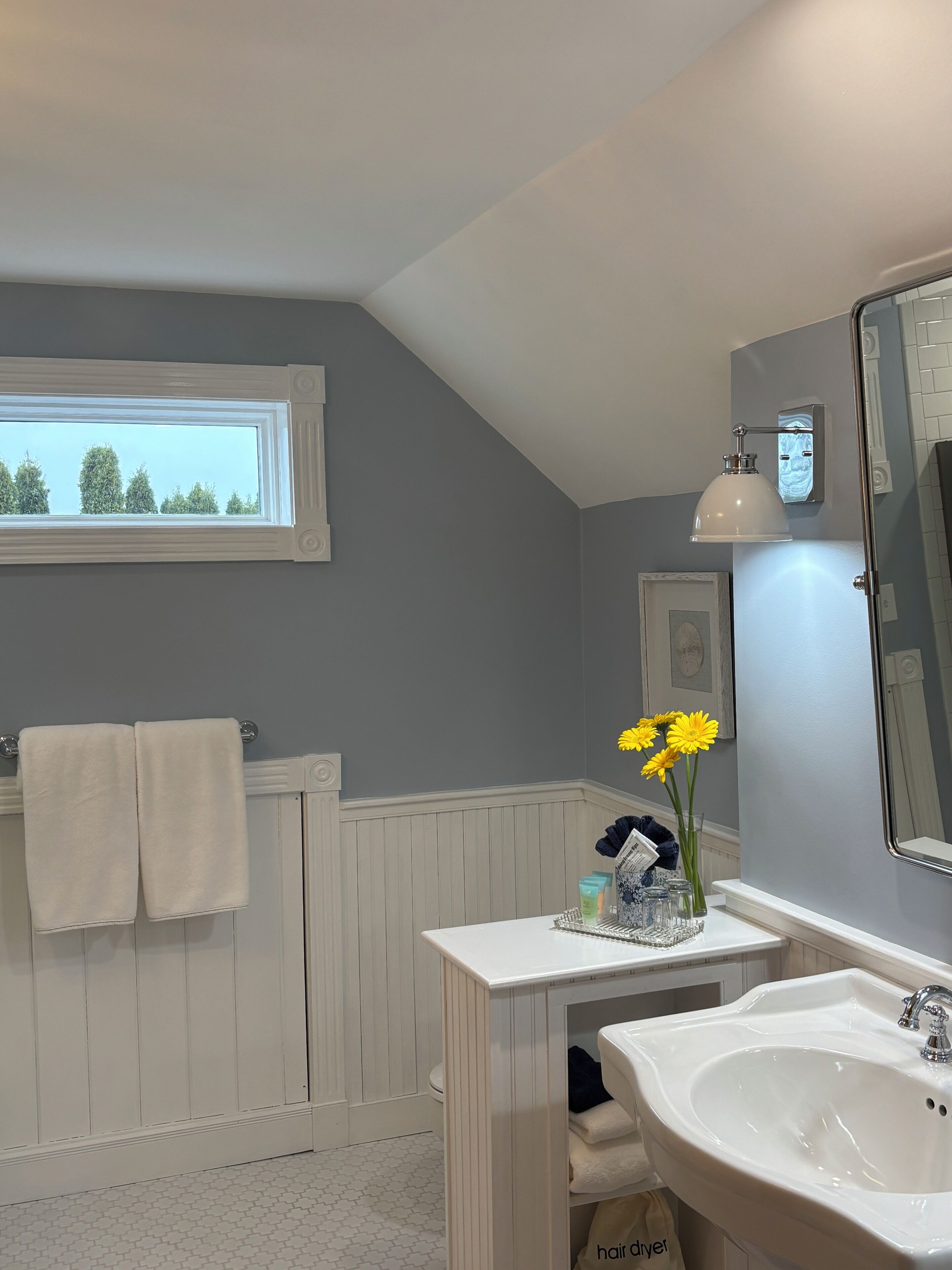 Une salle de bain avec un meuble-lavabo blanc, un grand miroir, une fenêtre donnant sur la verdure et un porte-serviettes avec deux serviettes.