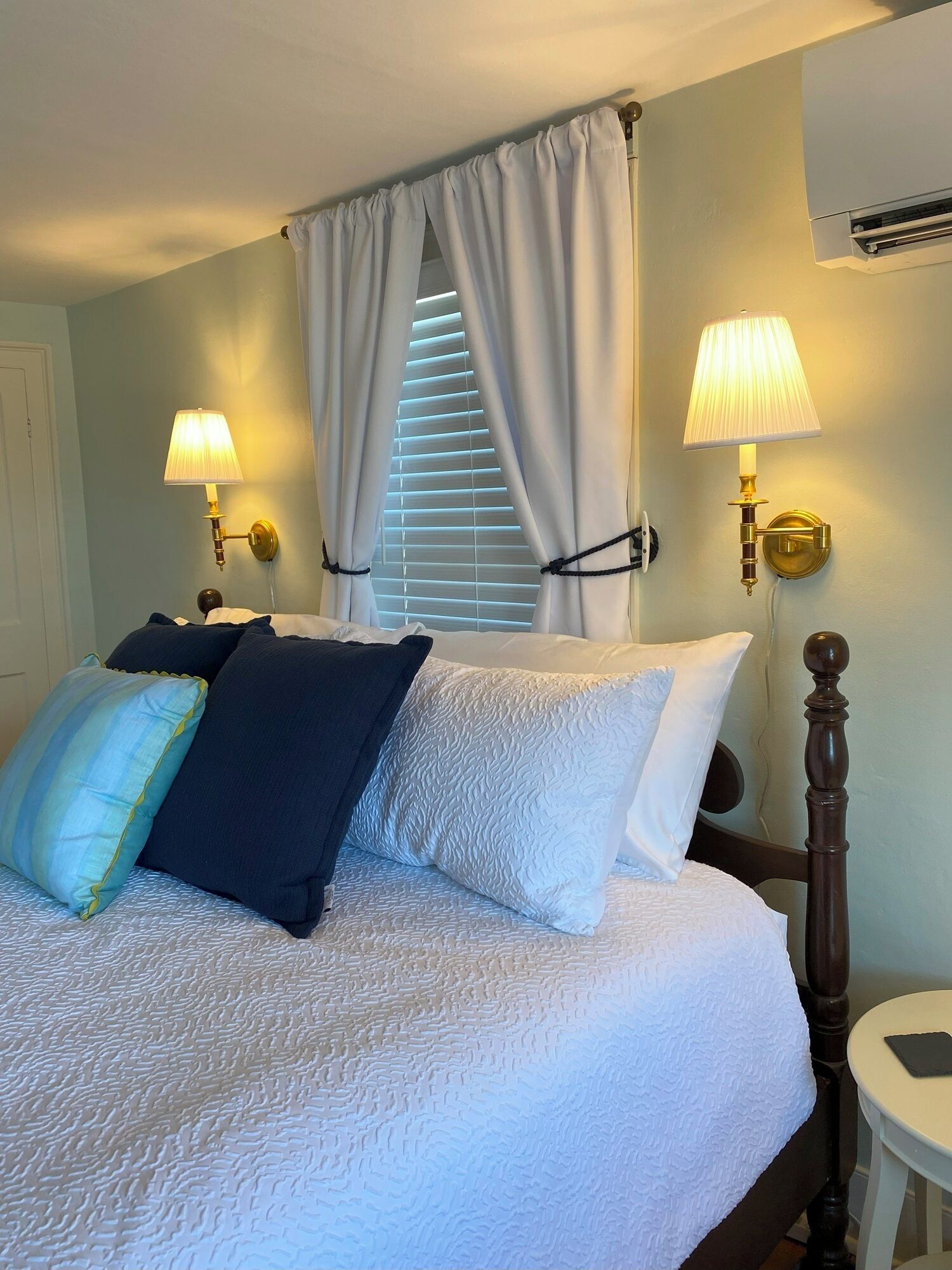 Una cama bien hecha con edredón blanco y varios almohadones, dos lámparas fijadas a la pared, una ventana con persianas y un equipo de aire acondicionado en la pared.