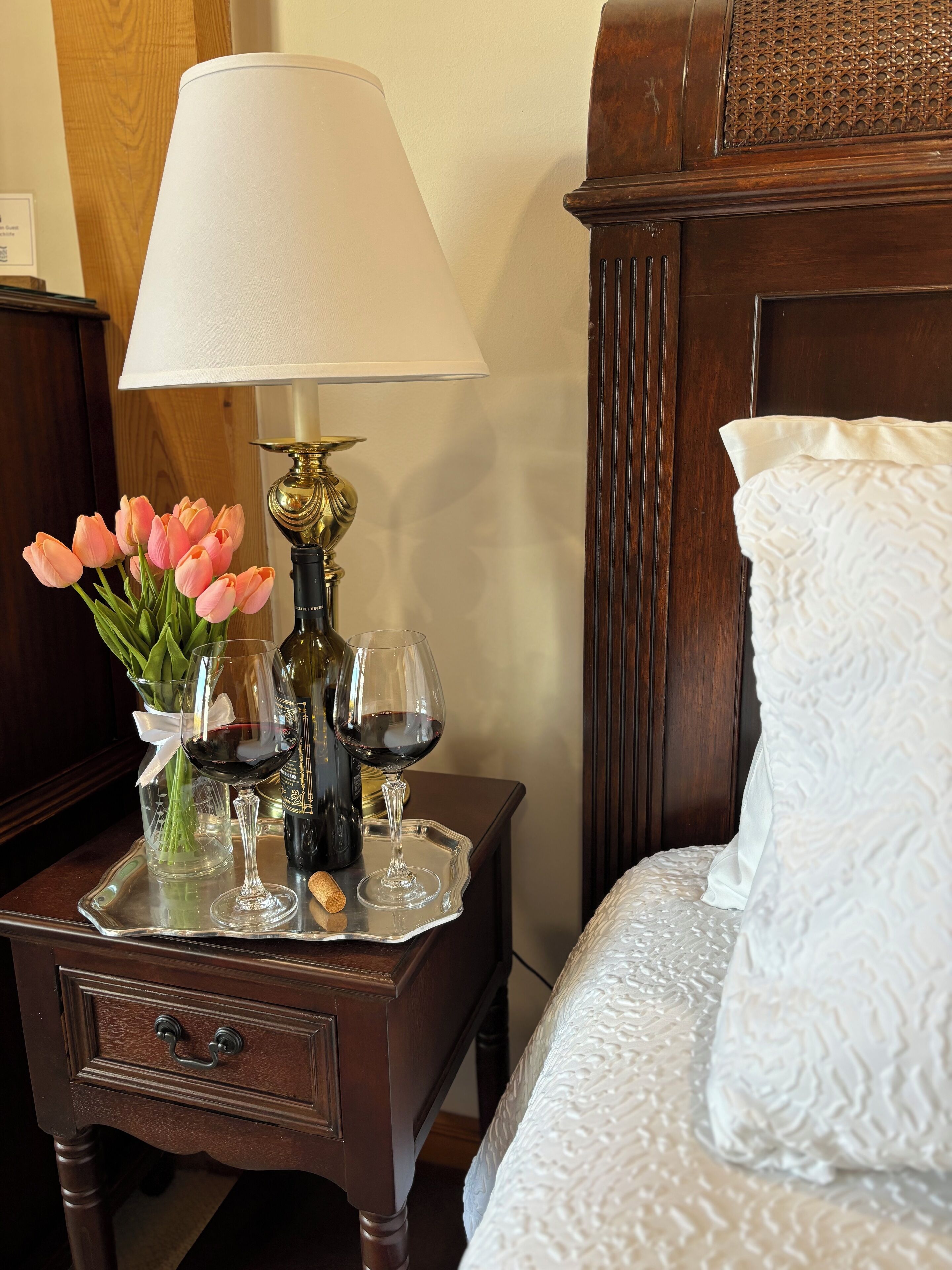 A wooden nightstand with a lamp, a tray holding wine glasses, a bottle, and a vase with pink tulips.