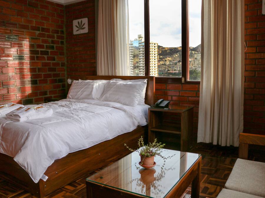 A hotel room with a brick wall, a bed with white linens, a bedside table with a phone, and a window with curtains.