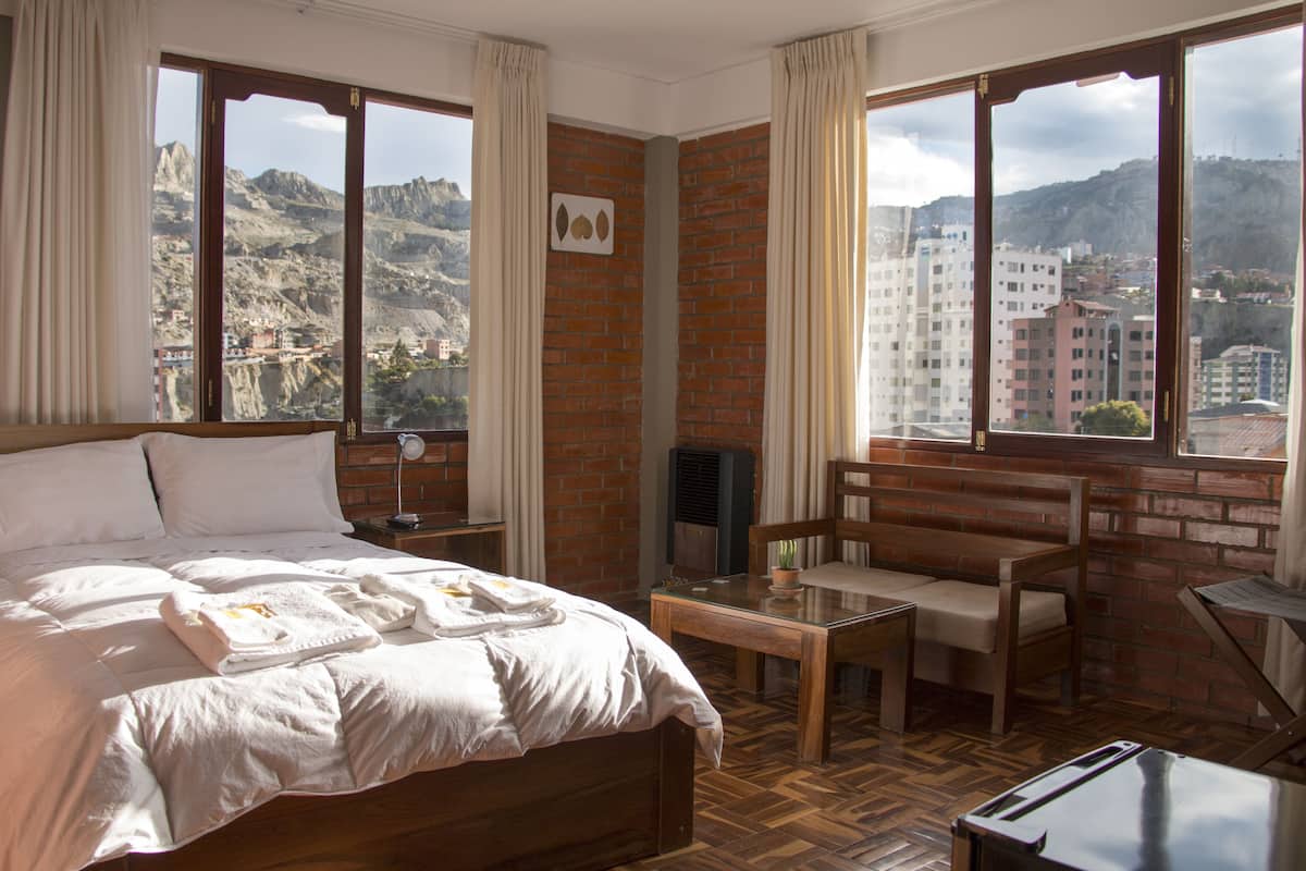 A hotel room with a bed, a small table, a chair, and a TV. The room has large windows with a view of a city and mountains.
