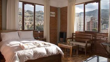 A hotel room with a bed, a small table, a chair, and a TV. The room has large windows with a view of a city and mountains.