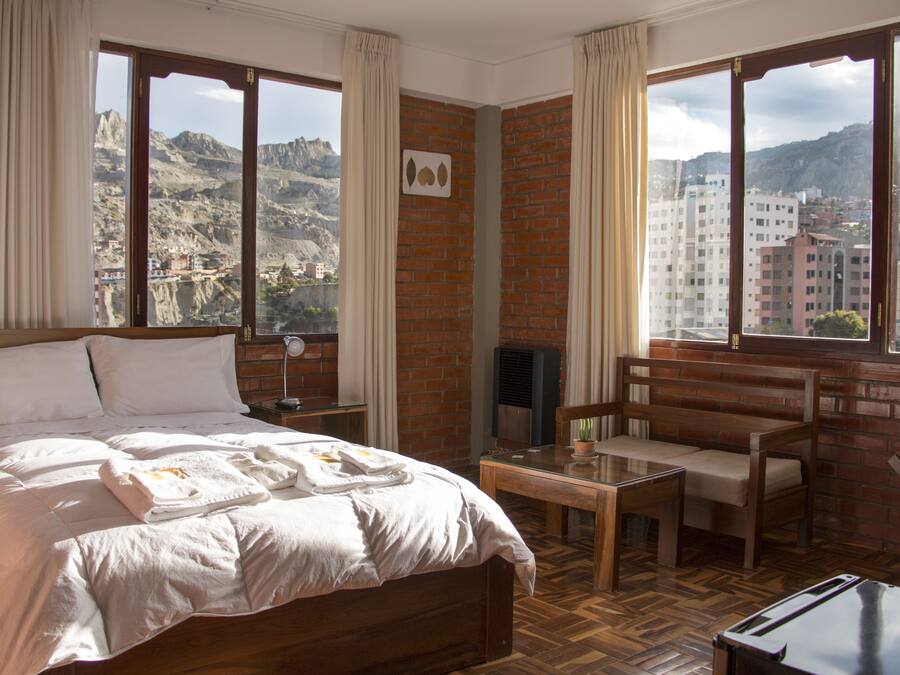 A hotel room with a bed, a small table, a chair, and a TV. The room has large windows with a view of a city and mountains.