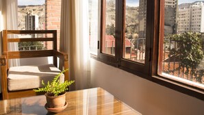 A room with a wooden chair, a small table, and a potted plant. A large window offers a view of buildings and hills. - Qhini Hotel Boutique (La Paz)