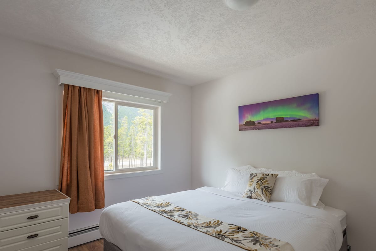 A bedroom with a bed, a dresser, a window with curtains, and a painting of the aurora borealis.