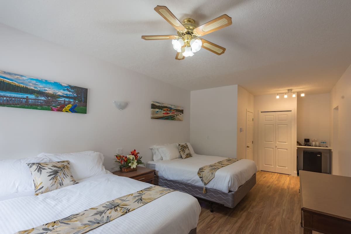 A hotel room with two beds, a ceiling fan, a painting of a landscape, a small table with a vase of flowers, and a door leading to a bathroom.
