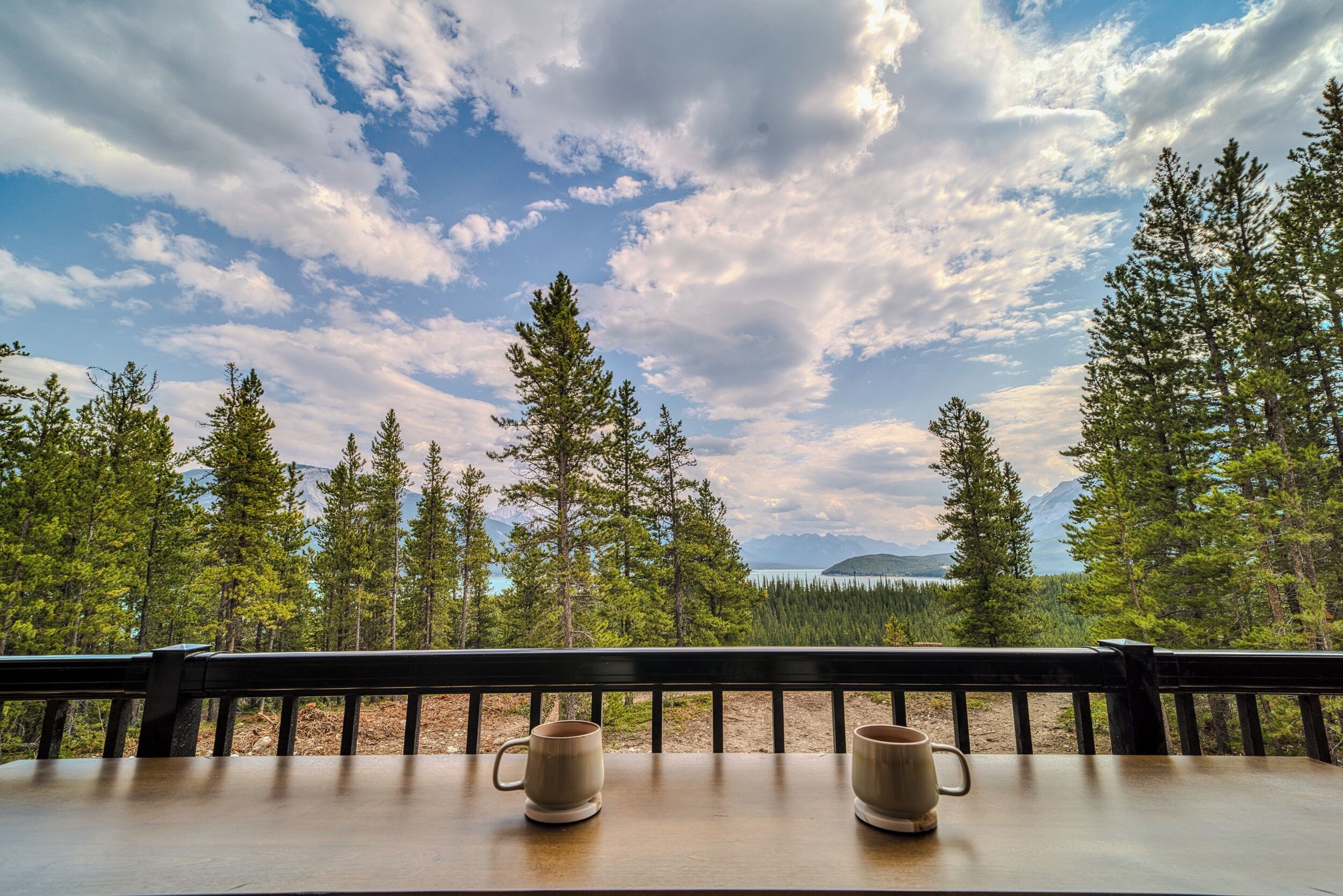 A wooden table with two empty ceramic mugs on a balcony overlooking a forest and mountains.