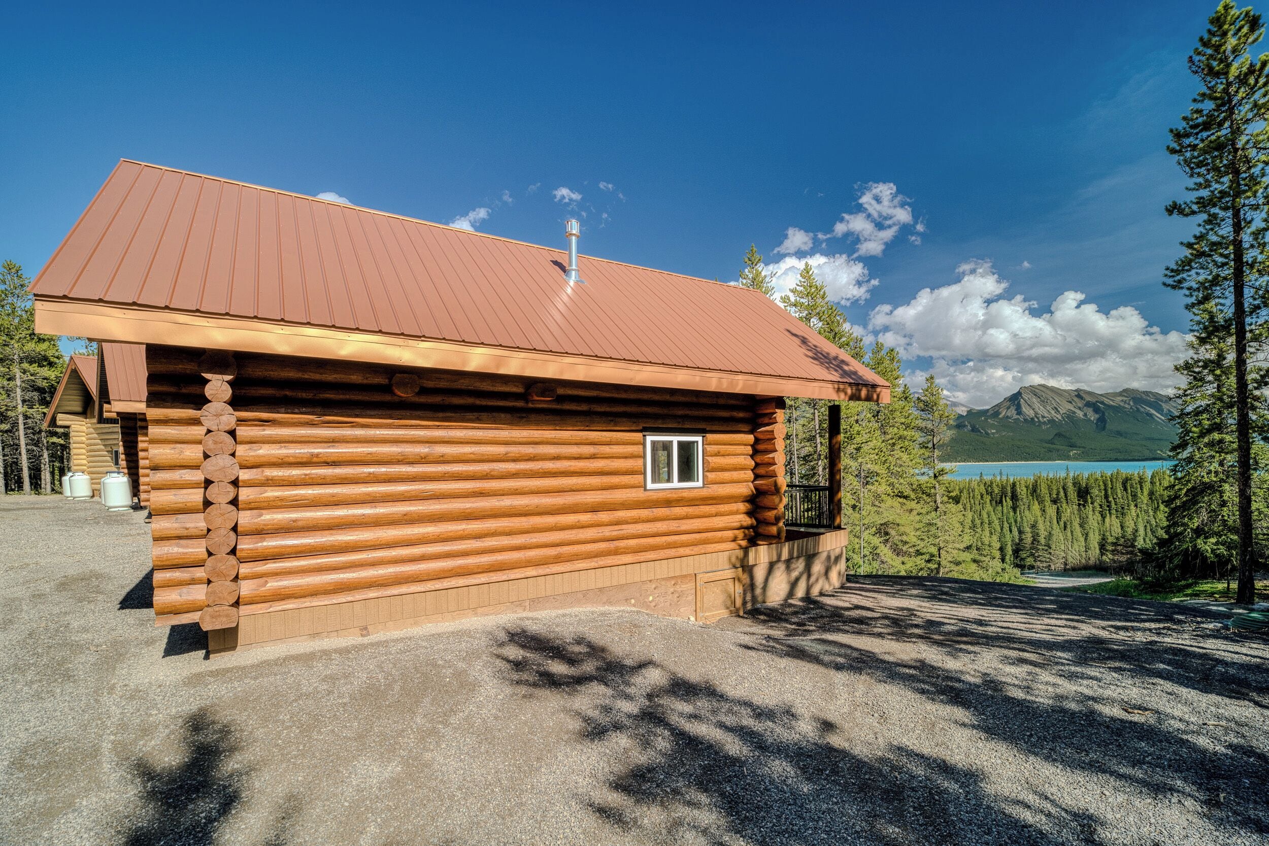 A log cabin with a metal roof, a small window, and a view of a lake and mountains in the background.