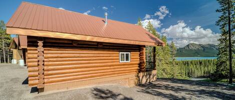 A log cabin with a metal roof, a small window, and a view of a lake and mountains in the background.