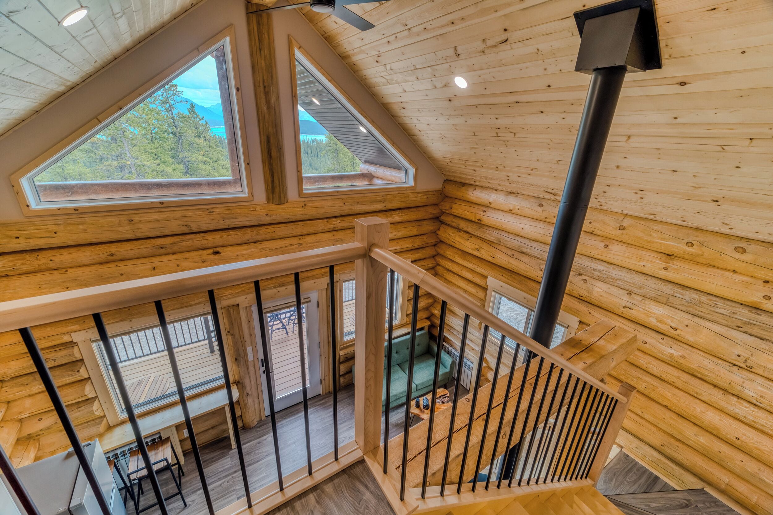 A wooden cabin interior with a loft bedroom, a staircase, and a view of the outdoors.