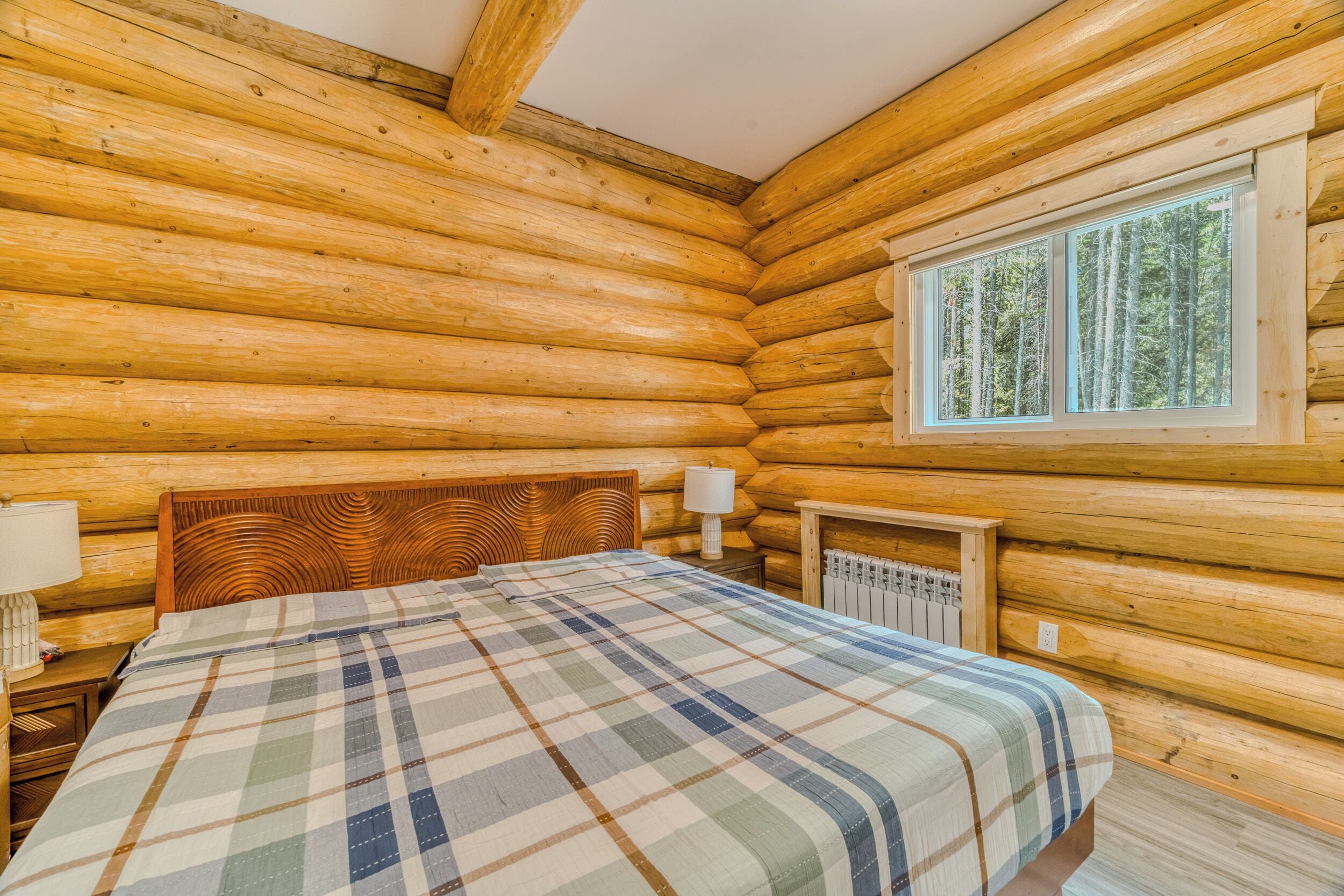 A log cabin room with a bed, a nightstand, a lamp, and a window with a view of trees.