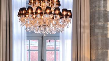 A living room with a chandelier, a sofa, a glass coffee table with flowers, a TV on a stand, and a view of a red brick building through a window.