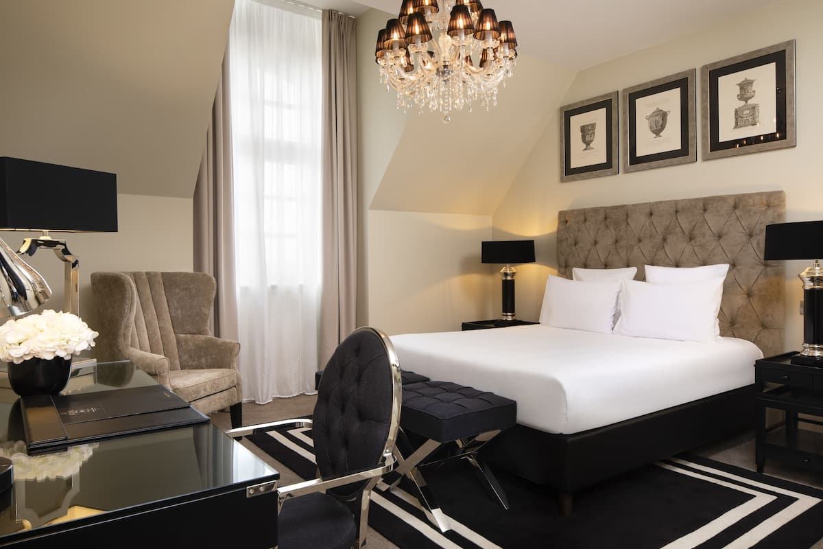 A hotel room with a large bed, a desk with a chair, a chandelier, framed pictures on the wall, and a window with curtains.