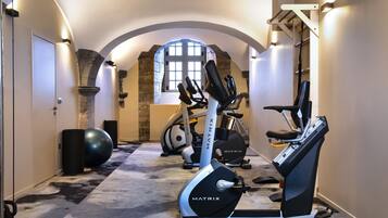A hallway with exercise equipment, including a treadmill and stationary bikes, in a room with arched ceilings and a large window.