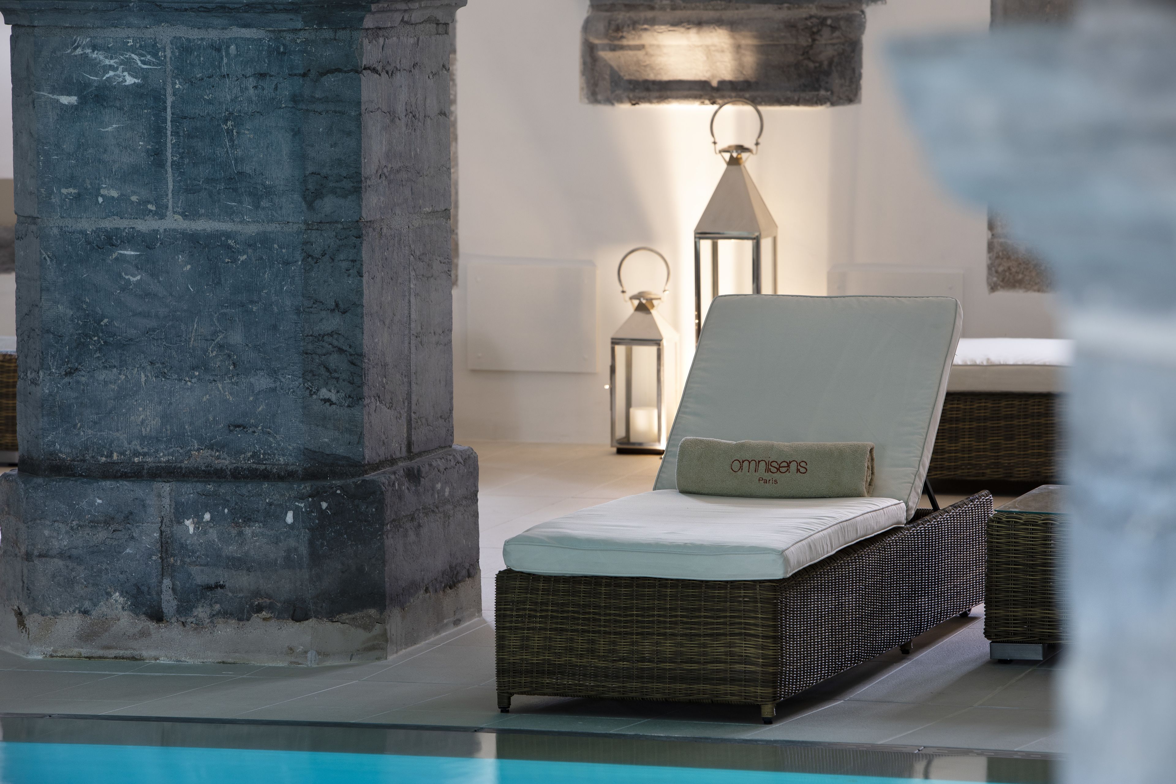 A lounge area with a wicker sofa, a cushion with "omnis Paris" text, a stone pillar, and a pool.