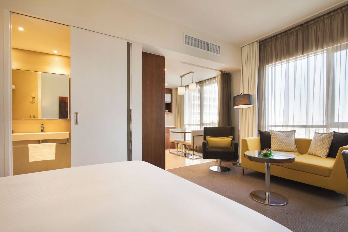 Quarto de hotel com uma cama grande, um sofá, uma mesinha, uma cadeira, banheiro com espelho e janela com cortinas.
