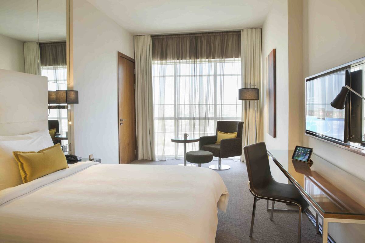 Quarto de hotel com uma cama grande, uma escrivaninha com computador, uma cadeira, uma mesinha e uma televisão.