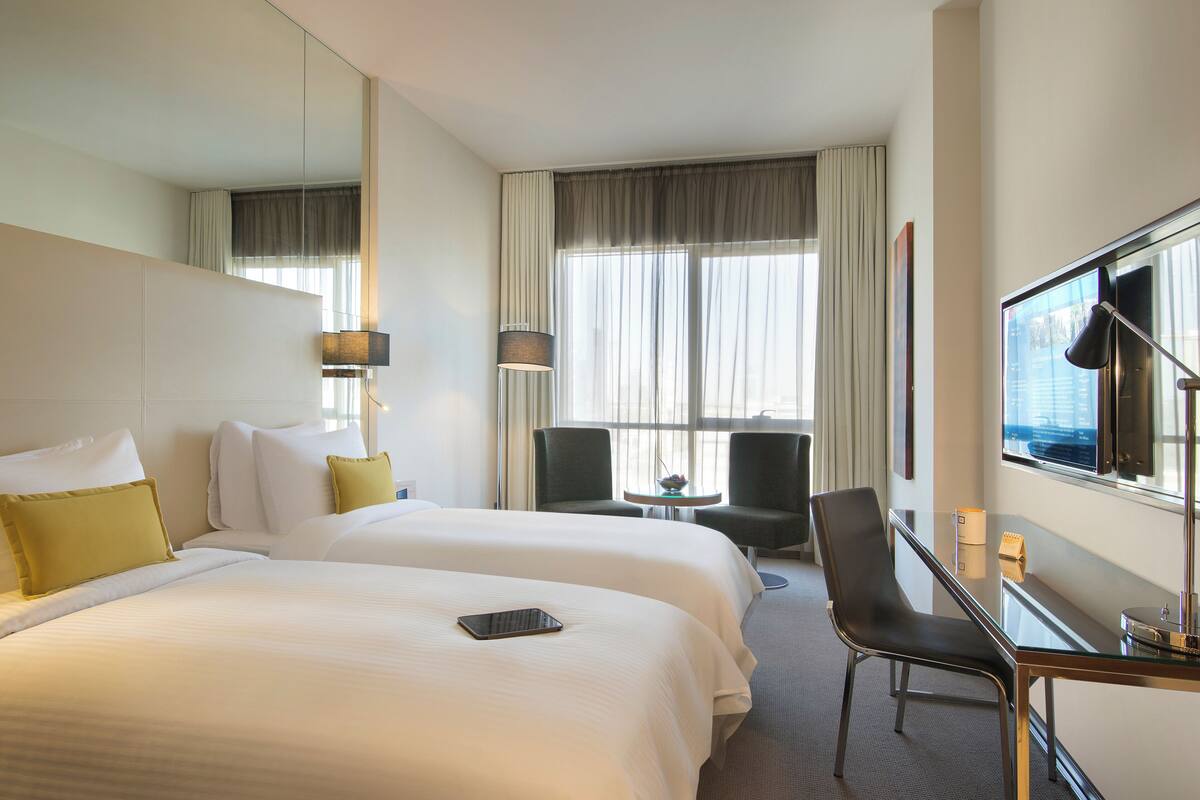 Quarto de hotel com duas camas, uma escrivaninha, uma cadeira, uma televisão e uma janela com cortinas.