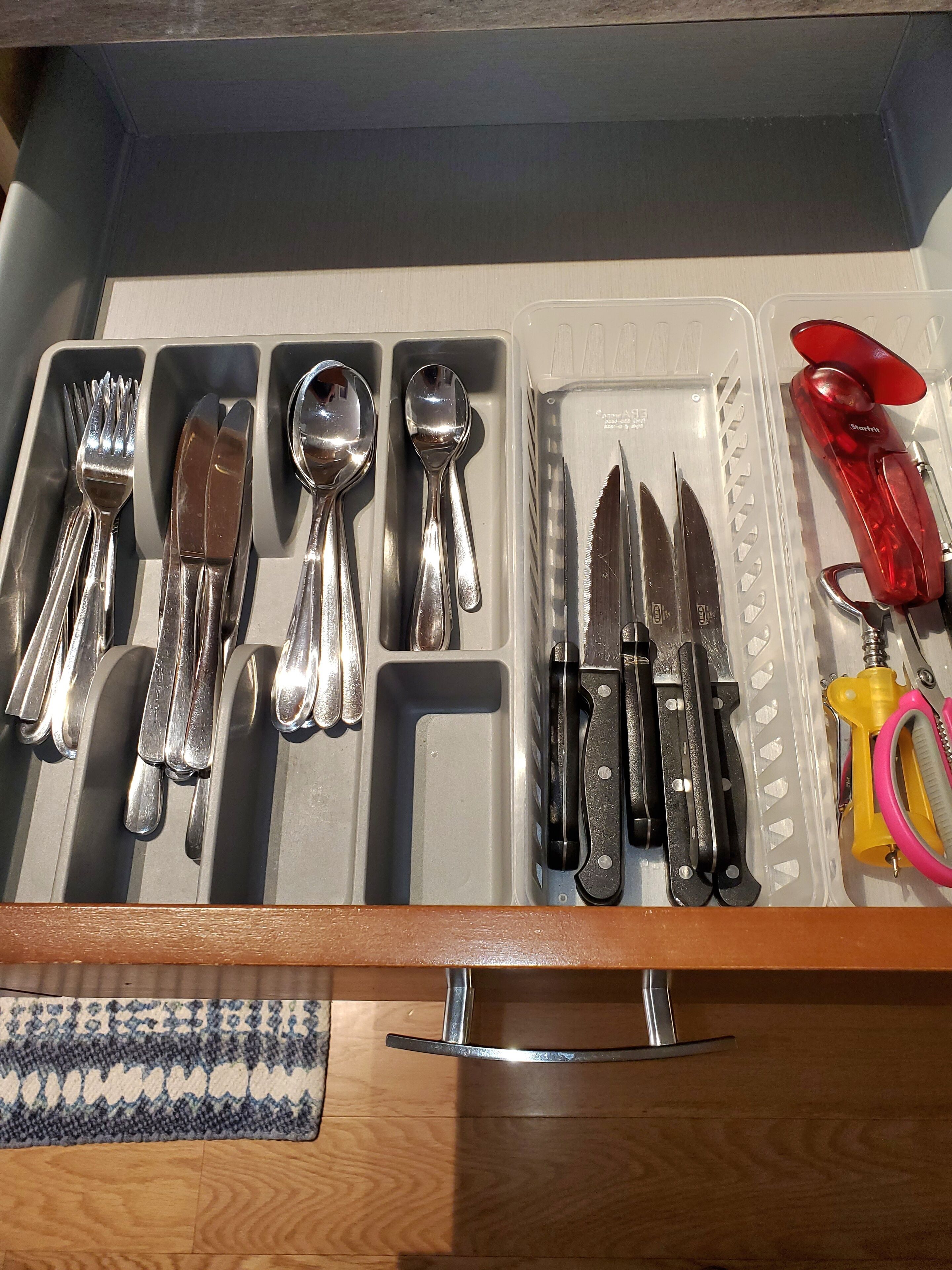 Un cajón con diversos utensilios y herramientas de cocina, como tenedores, cuchillos, cucharas y un afilador de cuchillos con mango rojo.