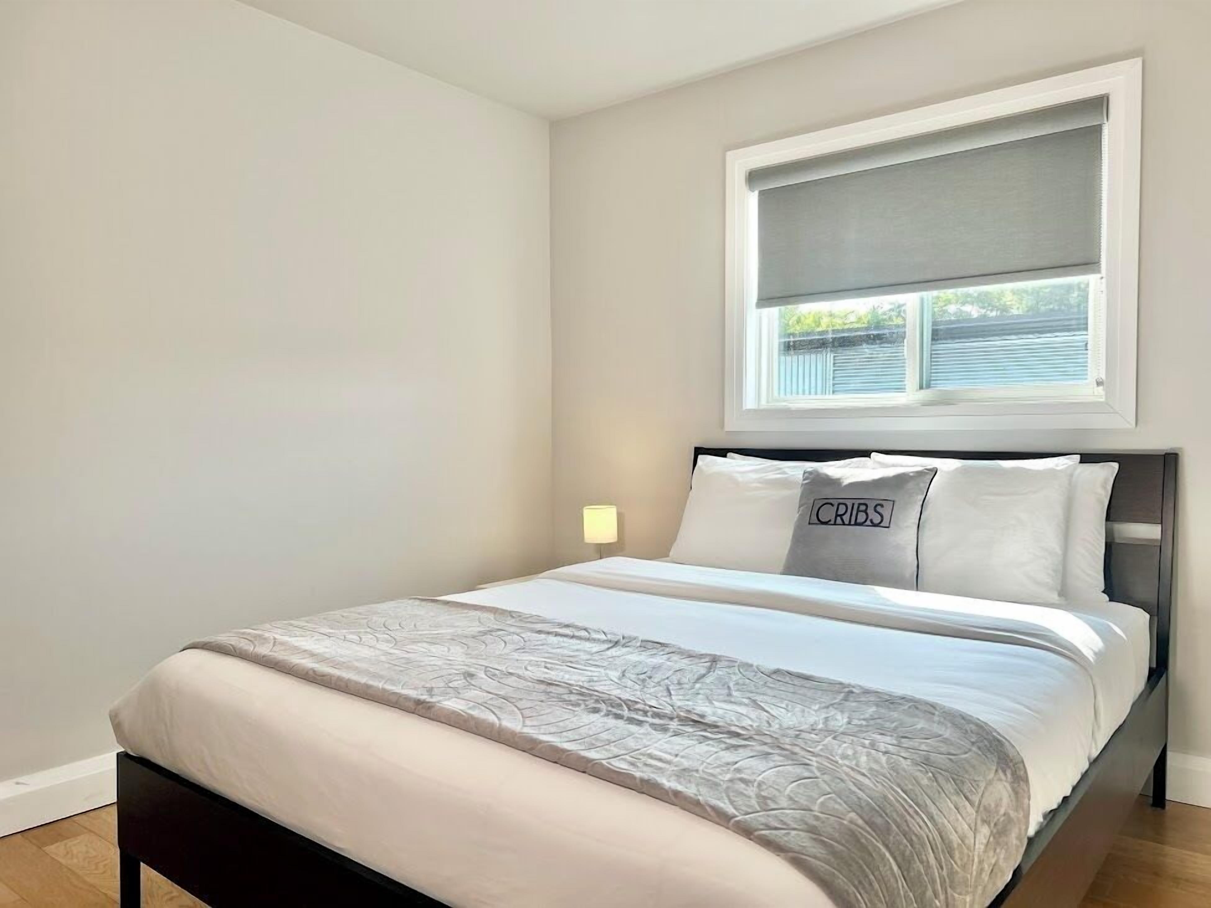 A bedroom with a bed, a window with blinds, and a wall-mounted lamp.