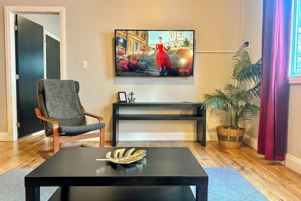 Ein Wohnzimmer mit an der Wand montiertem Fernseher, einem Holzstuhl, einem schwarzen Couchtisch und einer Topfpflanze.