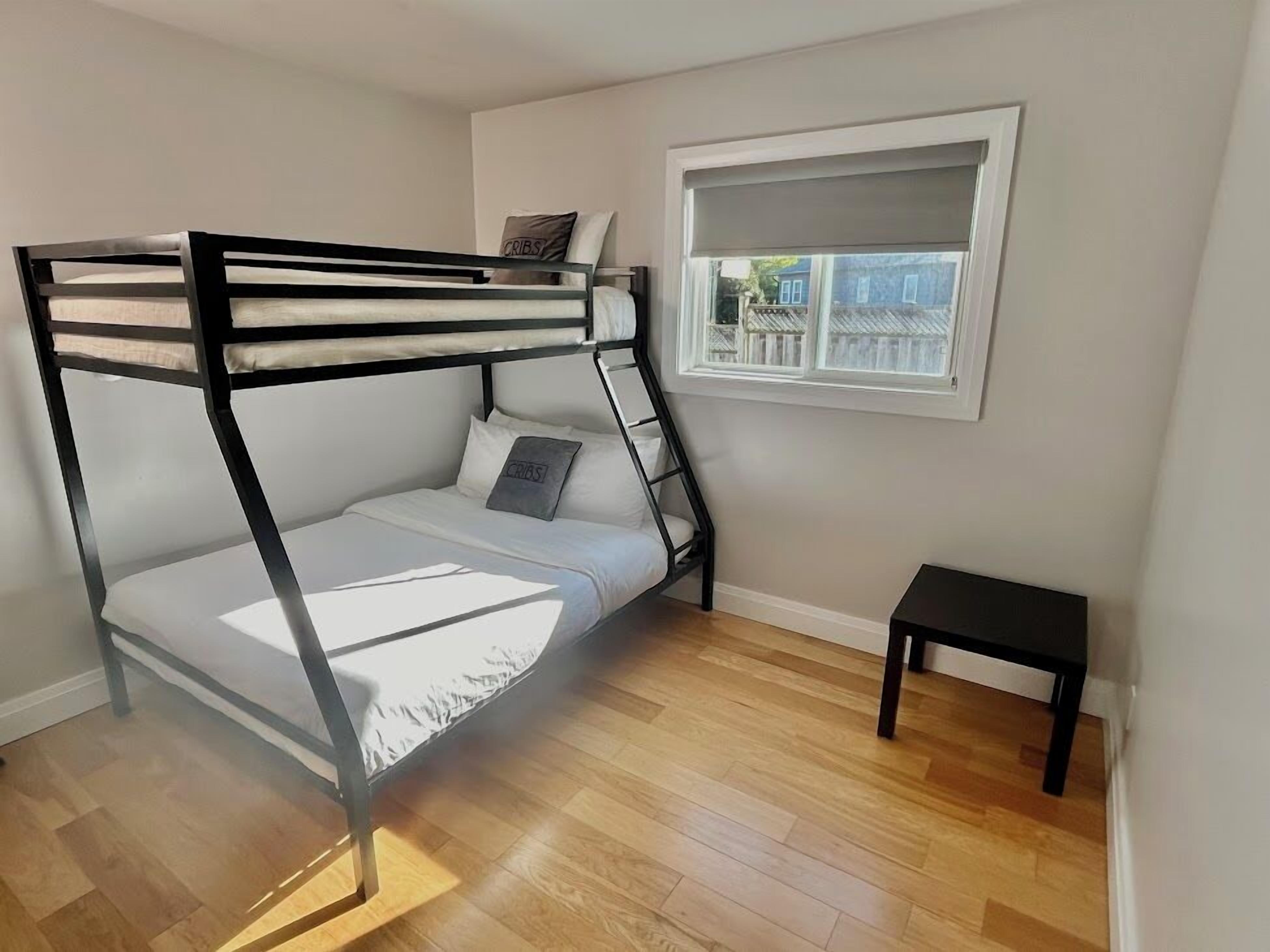 A bunk bed in a room with wooden flooring, a small table, and a window with a view of a fence.