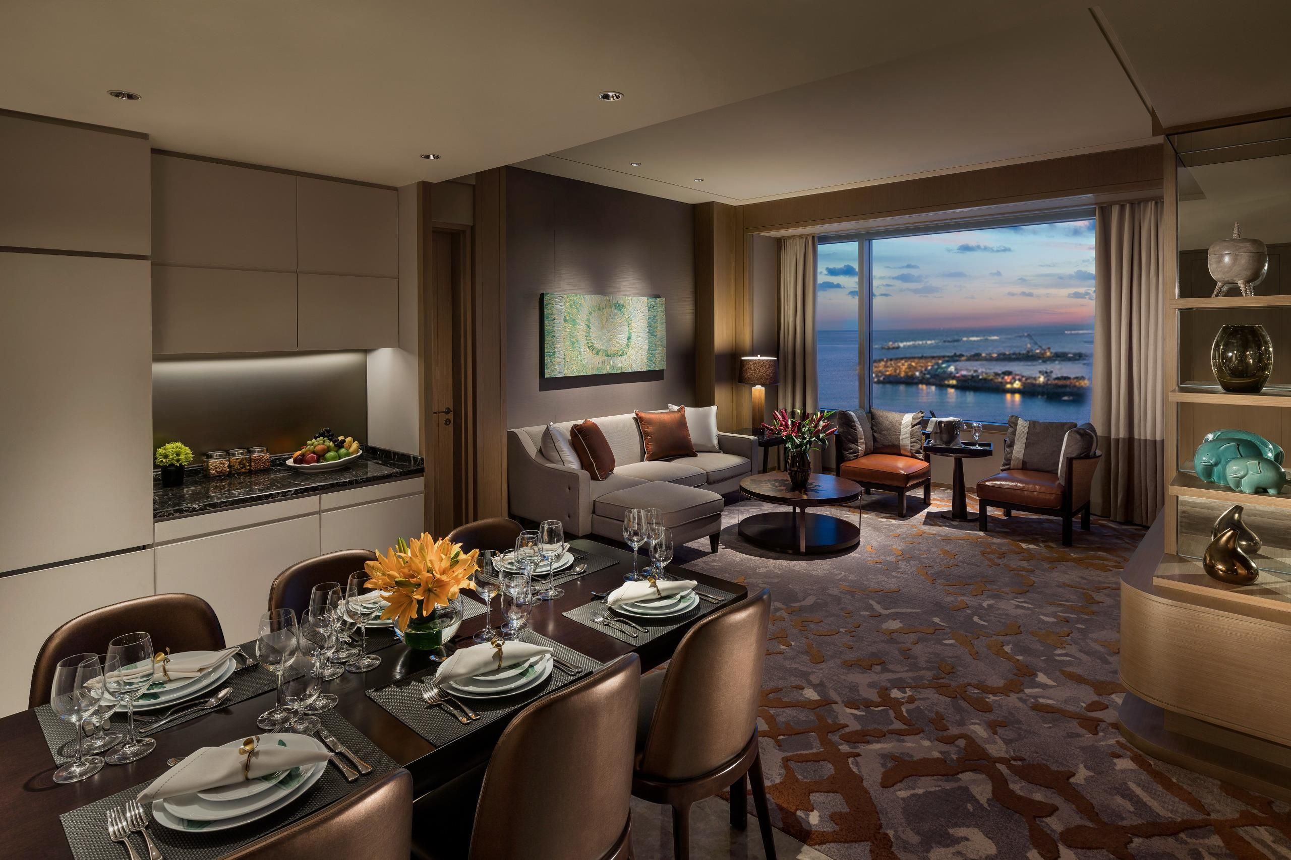 Habitación de hotel moderna con zona de comedor, sofá, mesa de centro y vistas a la ciudad.