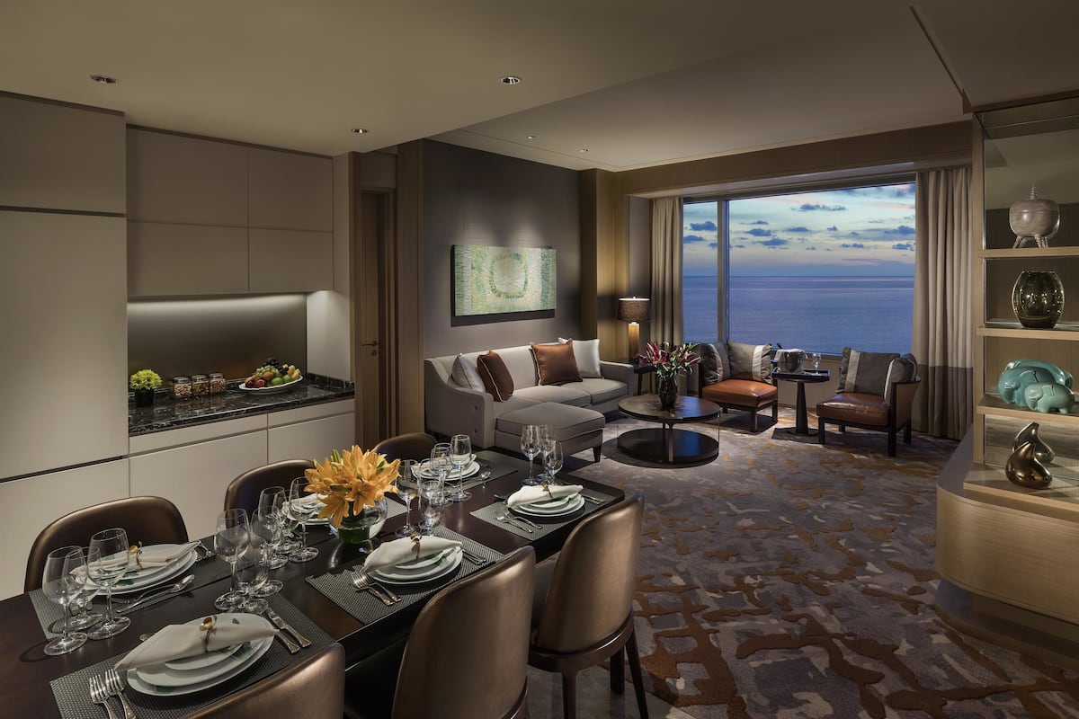 Une chambre d’hôtel moderne dotée d’un coin repas, d’un salon avec un canapé et des fauteuils, et d’une grande fenêtre offrant une vue sur la mer.
