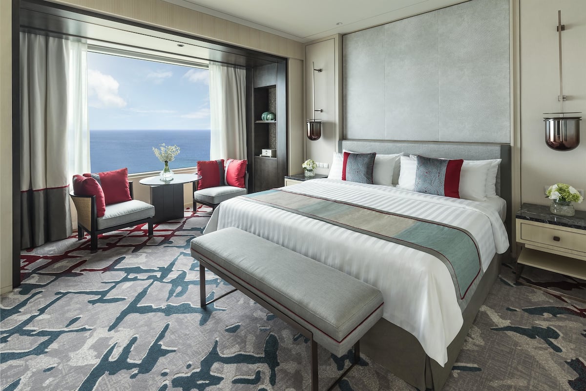 Une chambre d’hôtel avec un grand lit, un coin salon offrant une vue sur l’océan et une petite table sur laquelle est posé un vase de fleurs.