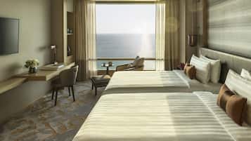 A hotel room with two beds, a desk, a chair, a TV, and a view of the ocean.