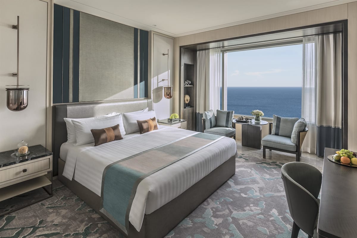 Une chambre d’hôtel moderne dotée d’un grand lit, d’un coin salon avec vue sur l’océan et d’une petite table sur laquelle est posé un panier de fruits.
