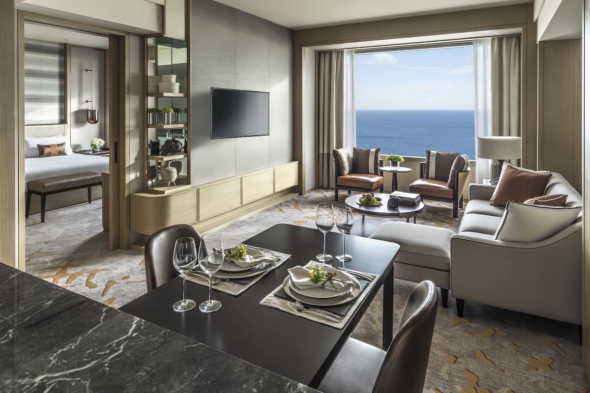 Une chambre d’hôtel moderne dotée d’un coin repas, d’un ensemble de canapés, d’une télévision à écran plat et d’une grande fenêtre offrant une vue sur l’océan.