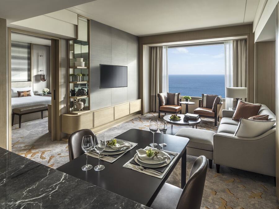 A modern hotel room with a dining area, a sofa set, a flat-screen TV, and a large window with a view of the ocean.