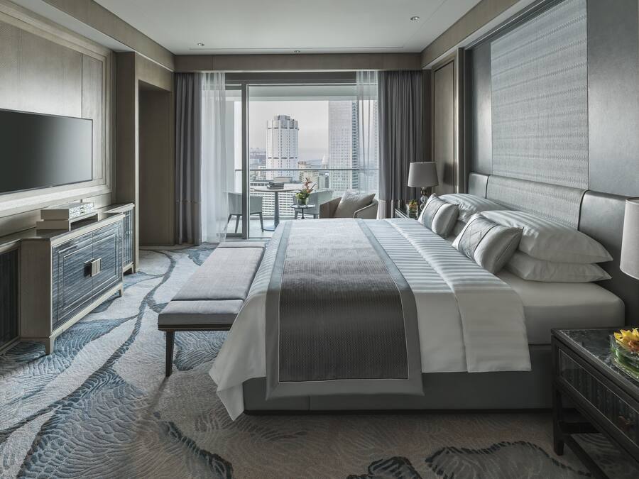 A modern hotel room with a large bed, a flat-screen TV, a desk with a lamp, and a view of the city skyline.