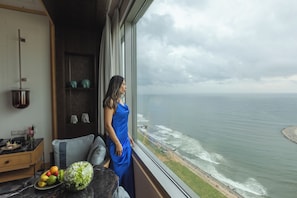 A woman in a blue dress standing by a window overlooking a beach with waves and a distant shoreline. - Shangri-La Colombo (Colombo)