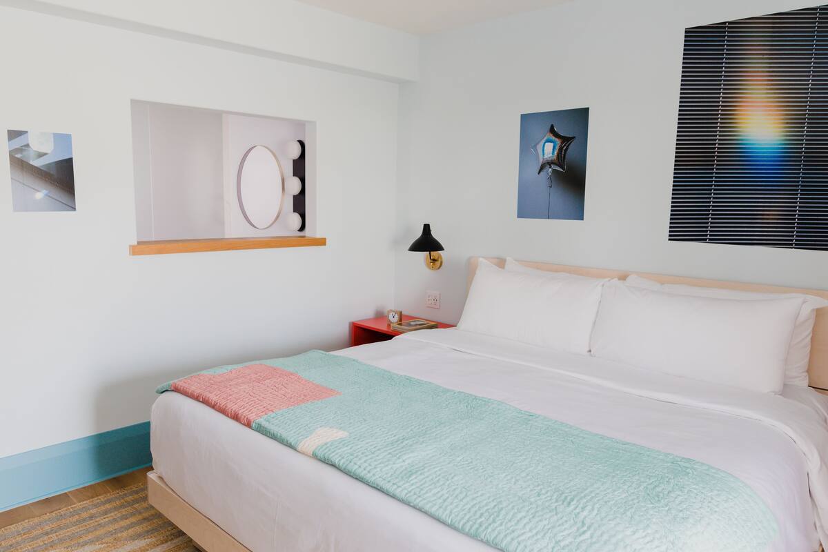 A neatly made bed with a light-colored duvet and a teal blanket, a small bedside table with a lamp, and a wall-mounted shelf with a mirror and lights.