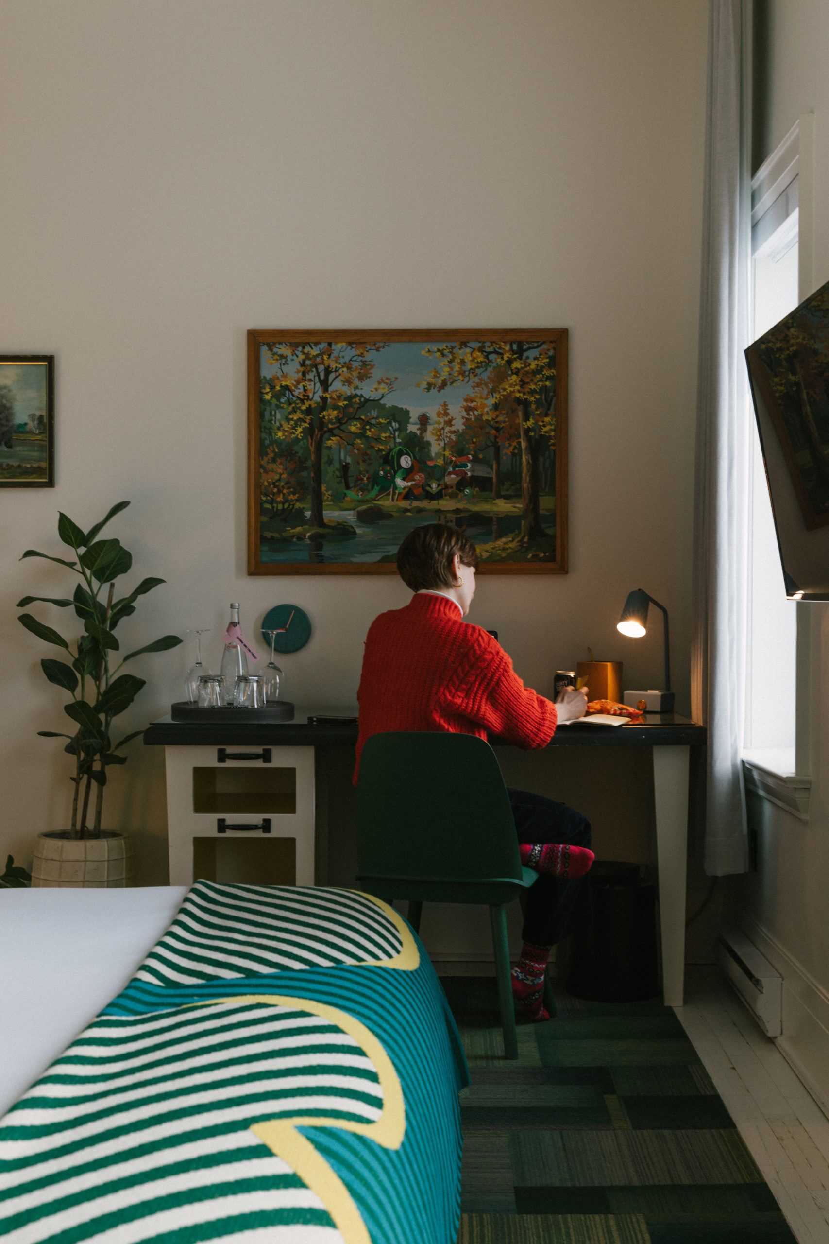 Ett hotellrum med en säng, ett skrivbord, en stol, en krukväxt, en tavla och ett fönster.