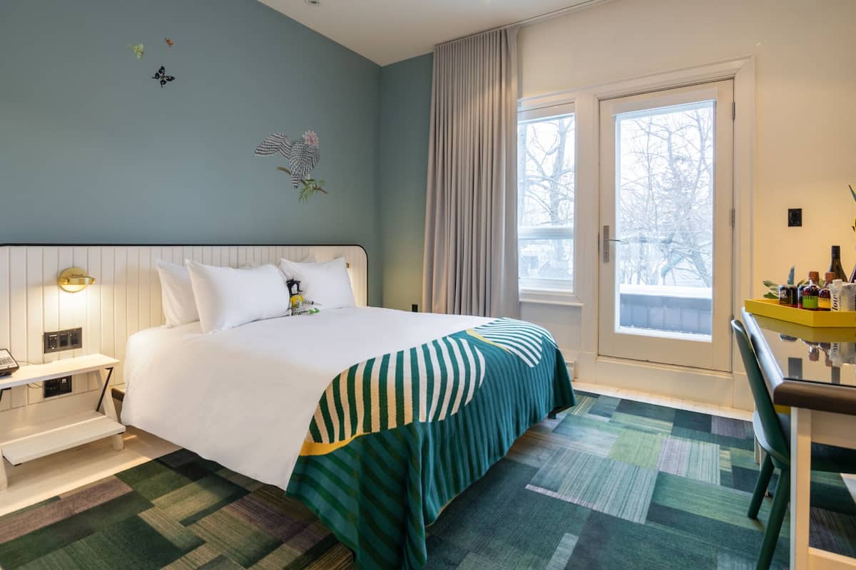 A hotel room with a bed, bedside table, and a chair. There is a window with curtains, a door, and a view of trees outside.