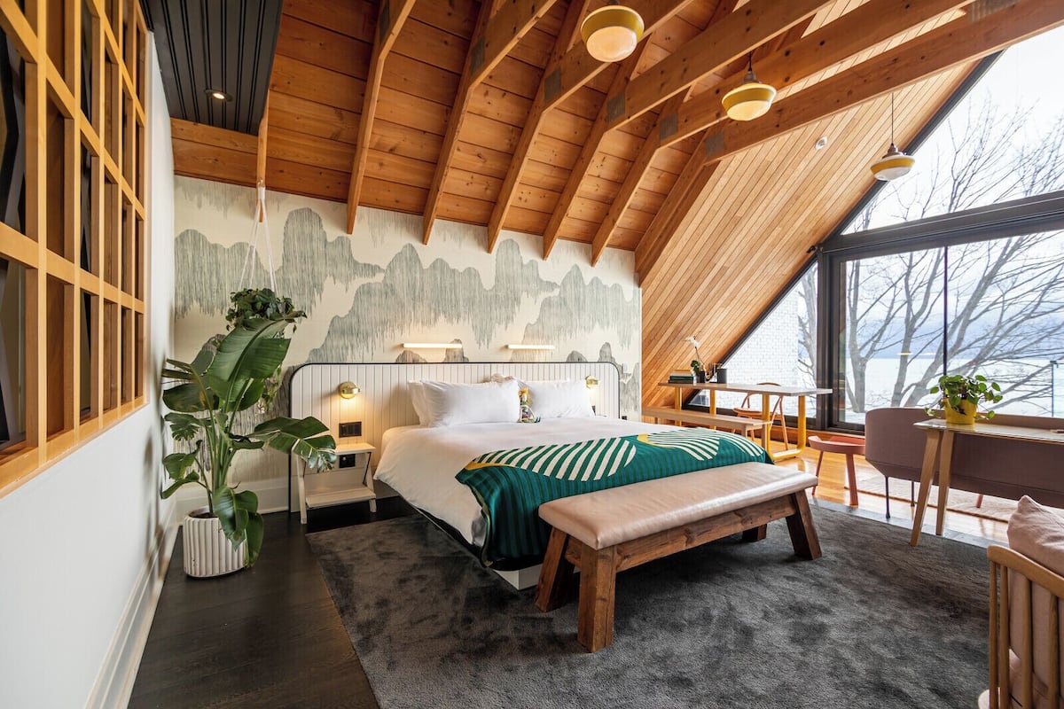 A bedroom with a bed, bedside tables, a bench, a desk, and a large window.