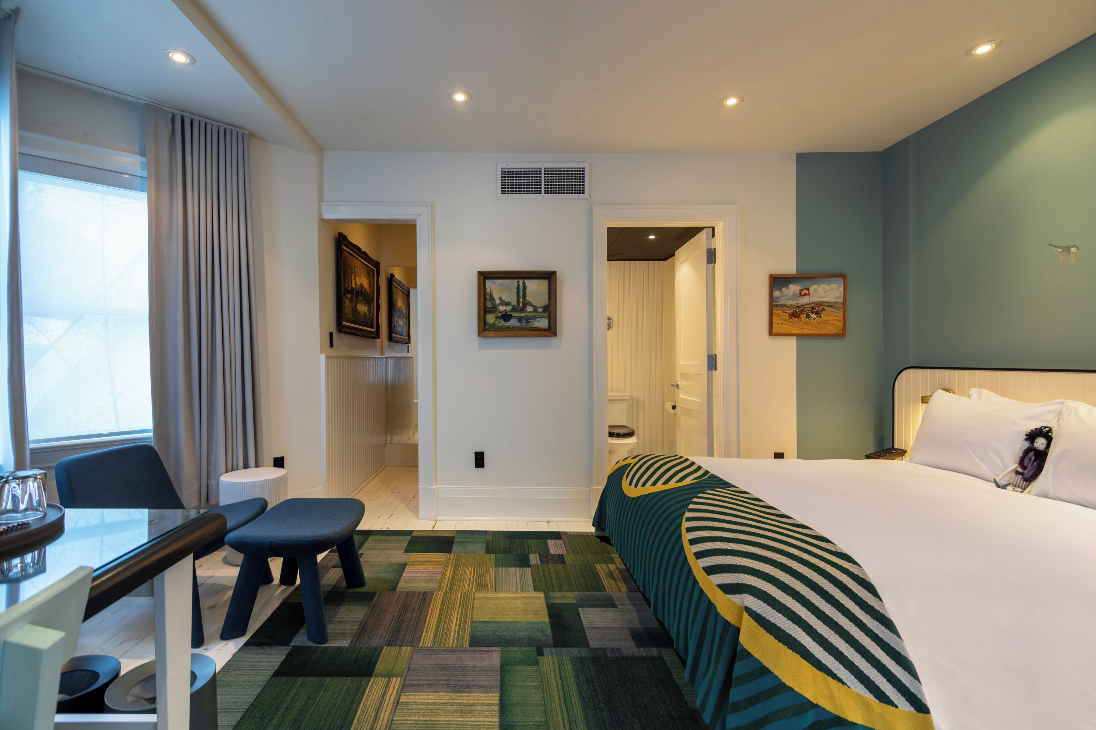 A hotel room with a bed, a desk, a chair, a bench, a radiator, a picture on the wall, and a door leading to another room.
