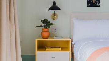 Ein ordentlich gemachtes Bett mit einem weißen Kopfkissen und einer pfirsichfarbenen Decke. Neben dem Bett steht ein kleiner gelber Nachttisch mit einer schwarzen Lampe und einem Topf mit einer Pflanze.