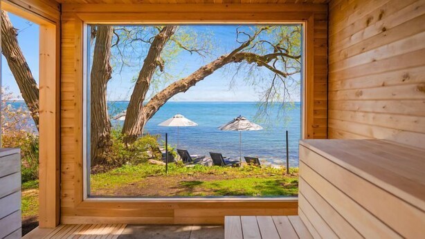 Una cabaña de madera con un ventanal grande con vistas a una playa con sillas de descanso y sombrillas.