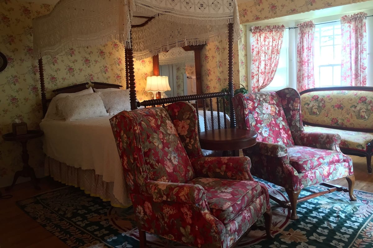 A four-poster canopy bed with floral wallpaper, a round wooden table, and floral armchairs.