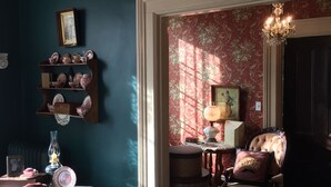 A room with a dark blue wall, a wooden chair with a cushion, a table with a lace tablecloth, and a patterned rug. - Annie Wiggins Guest House (Galena)