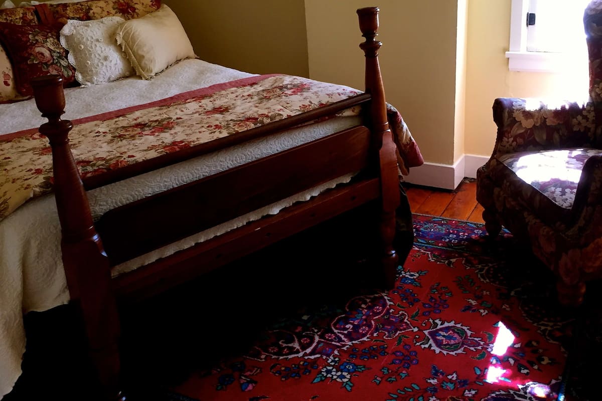 A bedroom with a wooden bed, a floral-patterned bedspread, a red patterned rug, and a chair with a floral design.