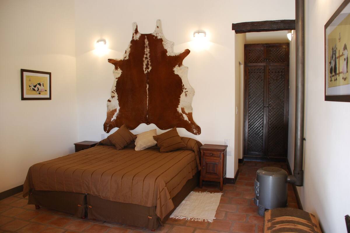 A bedroom with a large, brown and white animal hide headboard, a bed with brown bedding, a small wooden nightstand, a trash can, and a framed picture on the wall.