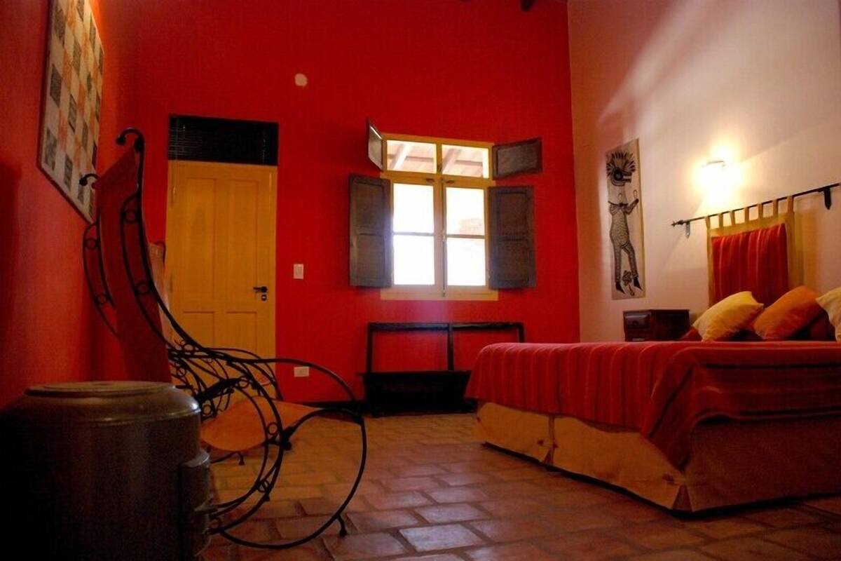 A room with a red wall, a bed with a red blanket, a chair, a window with shutters, and a wall-mounted light fixture.