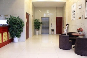 A hotel lobby with a reception desk, potted plants, an elevator, and seating areas.