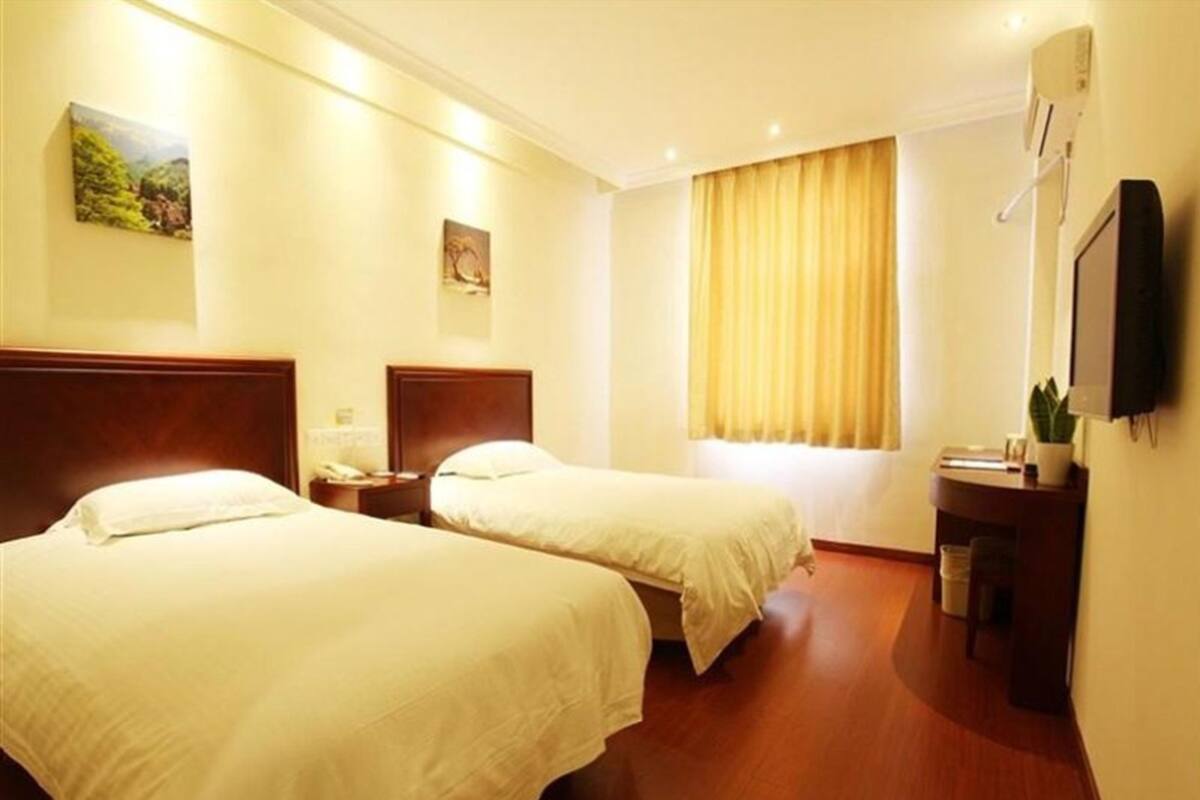 A hotel room with two single beds, a bedside table with a lamp, a small desk with a plant, and a window with curtains.