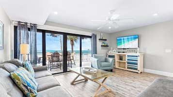 Una sala de estar con vista a la playa, un ventilador de techo, un televisor y una mesa de centro de madera.